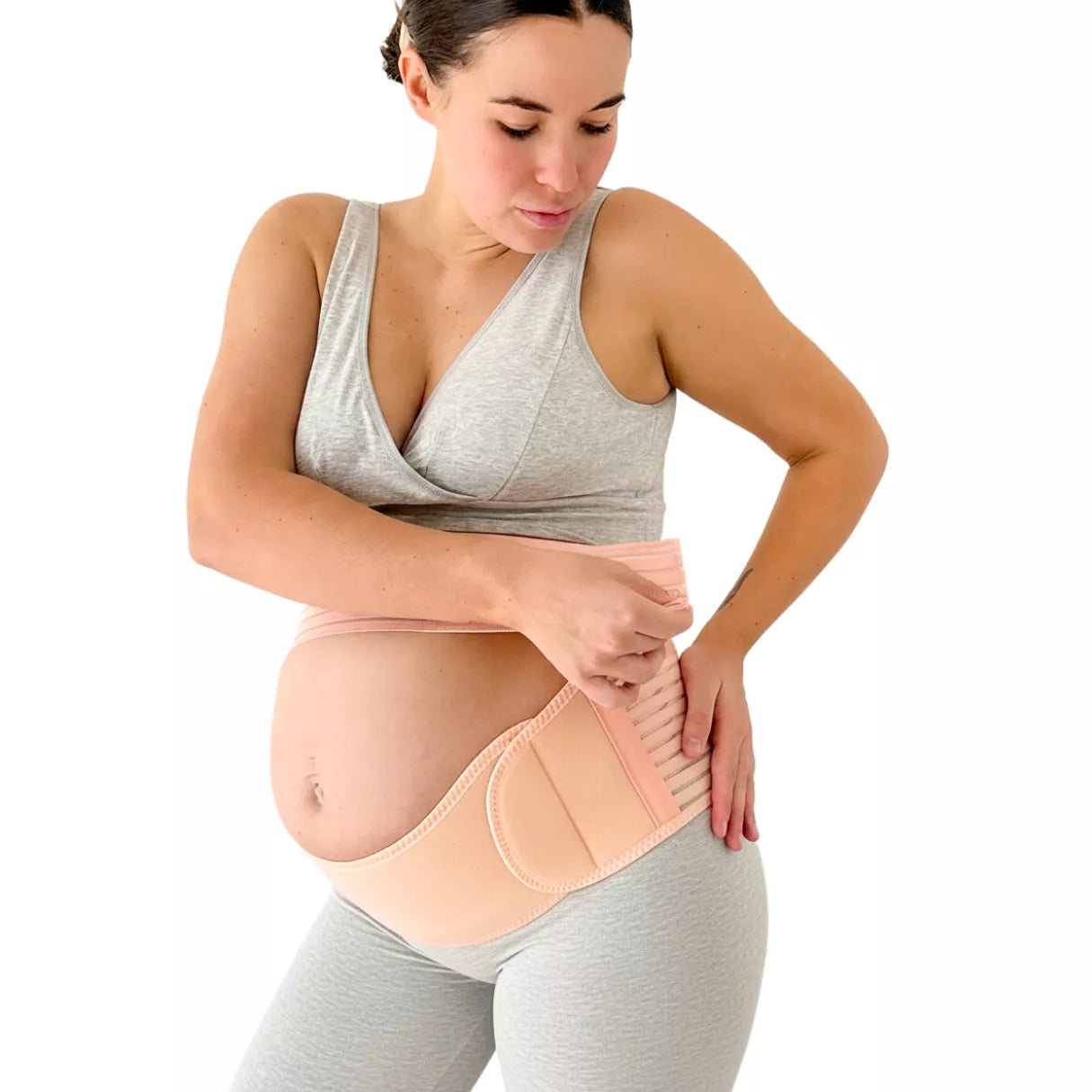 A Pregnant woman adjusting a beige pregnancy support brace band worn under the belly on a white background.