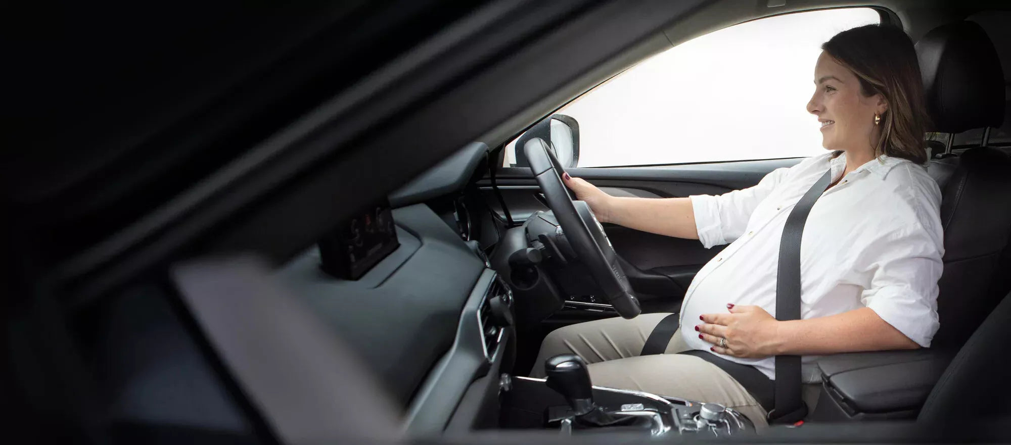 A pregnant woman driving a car using a seat belt clip to reposition the lap belt under the belly on a black background.